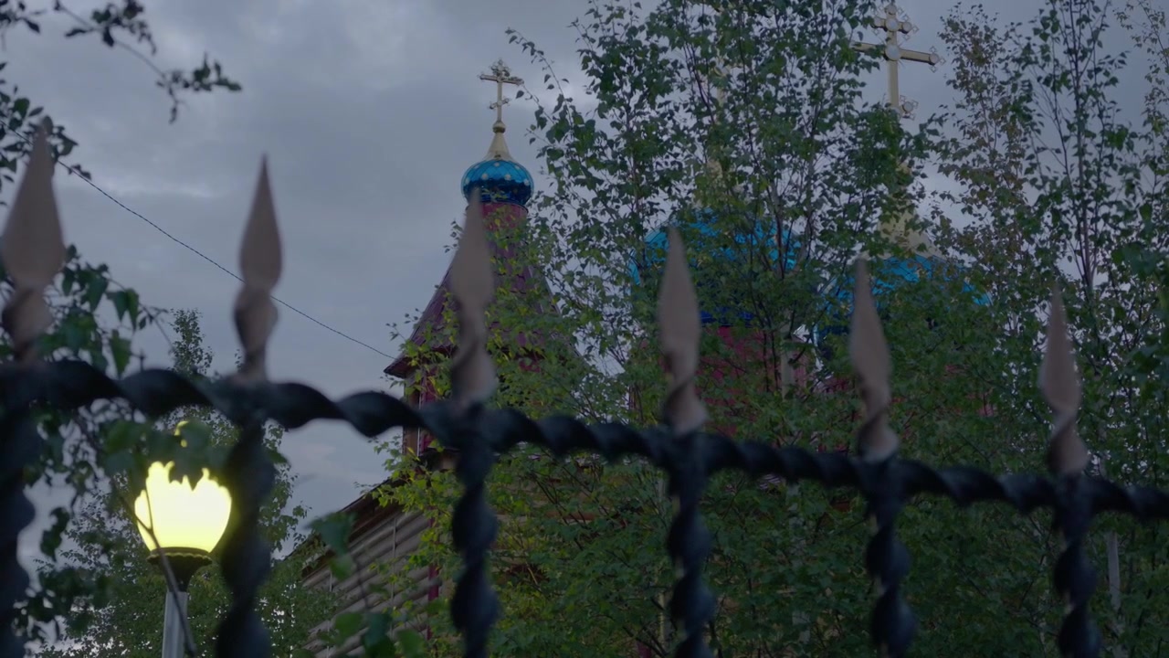 A static view through a black wrought-iron fence reveals a wooden church with blue domes and golden crosses, partially obscured by green foliage under an overcast sky.