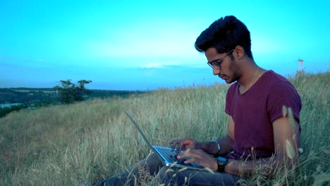 un tipo indio con gafas trabaja detrás de una computadora portátil sentado en la hierba en la cima de la colina sobre el río
