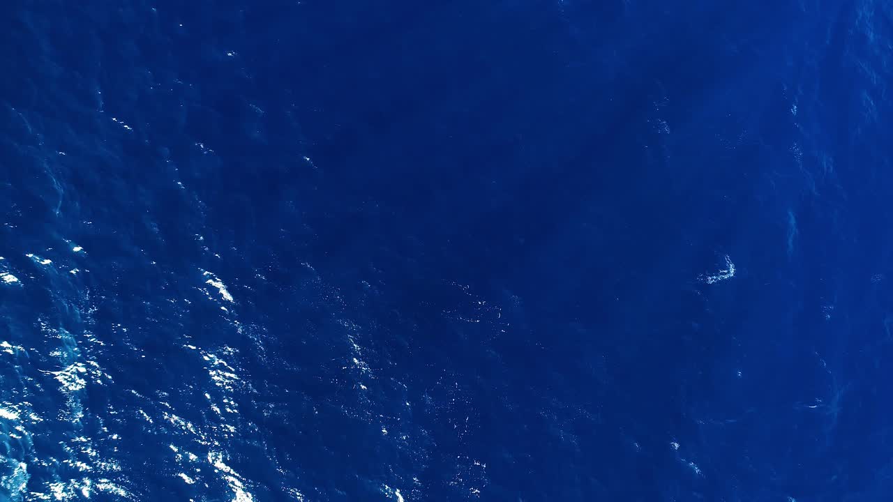 Drone stijgt verticaal op boven oceaanwater met diepblauwe textuurrimpelingen, zonlicht op golfpieken