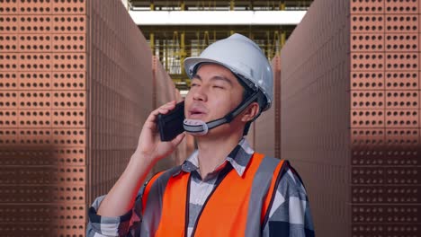 close up side view of asian male engineer with safety helmet talking on smartphone while standing with red brick packed in stacks are stored