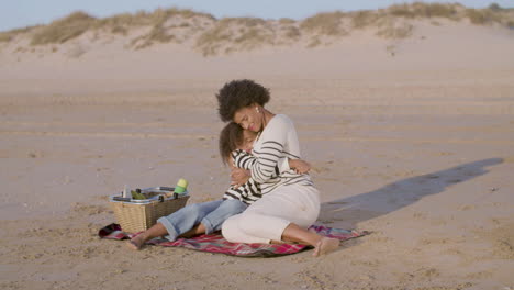 Happy-Mother-And-Daughter-Hugging-While-Having-Picnic-On-The-Beach-At-The-Sunset