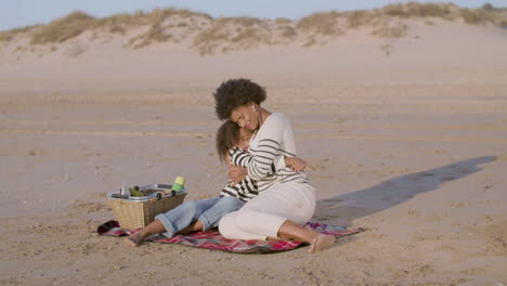 happy mother and daughter hugging while having picnic on the beach at the sunset