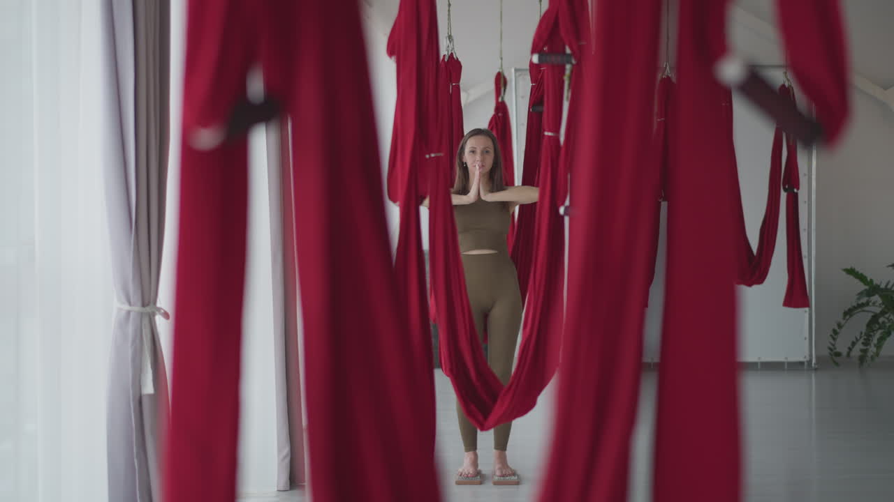Flexible lady practices aerial yoga between red silk hammocks lined up in bright studio balancing on nail board with hands in prayer pose focusing on strength flexibility mindfulness indoor