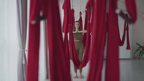 flexible lady practices aerial yoga between red silk hammocks lined up in bright studio balancing on nail board with hands in prayer pose focusing on strength flexibility mindfulness indoor