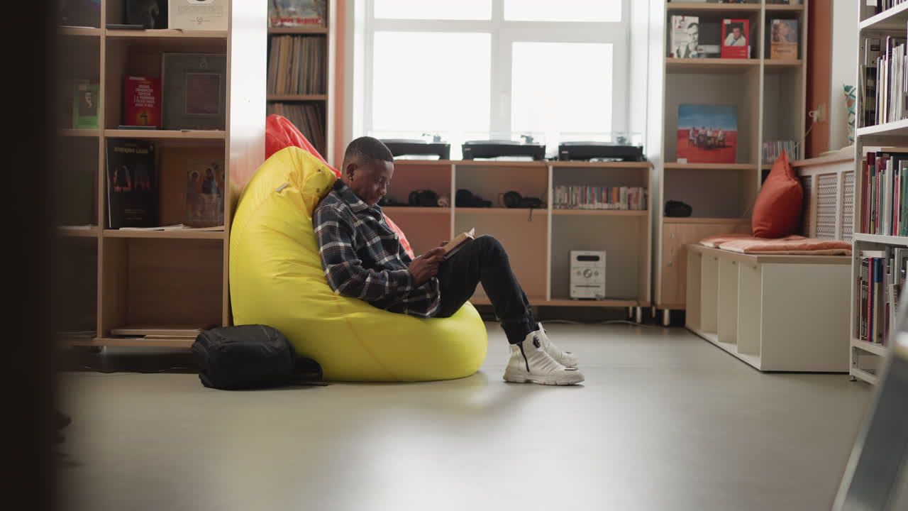 el hombre negro disfruta del libro sentado en una bolsa de frijoles. el lector masculino se sienta cómodamente en una silla acolchada en el salón de la biblioteca de la universidad absorto en el libro. lugar acogedor para la educación
