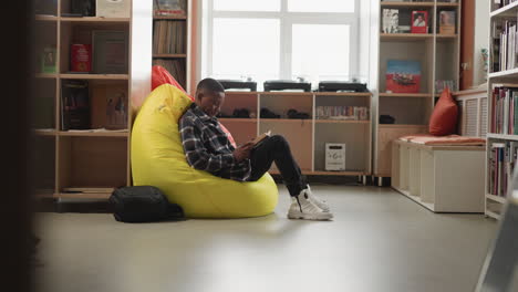Black man enjoys book sitting on bean bag. Male reader comfortably sits in cushioned chair in college library lounge engrossed in book. Cozy place for education