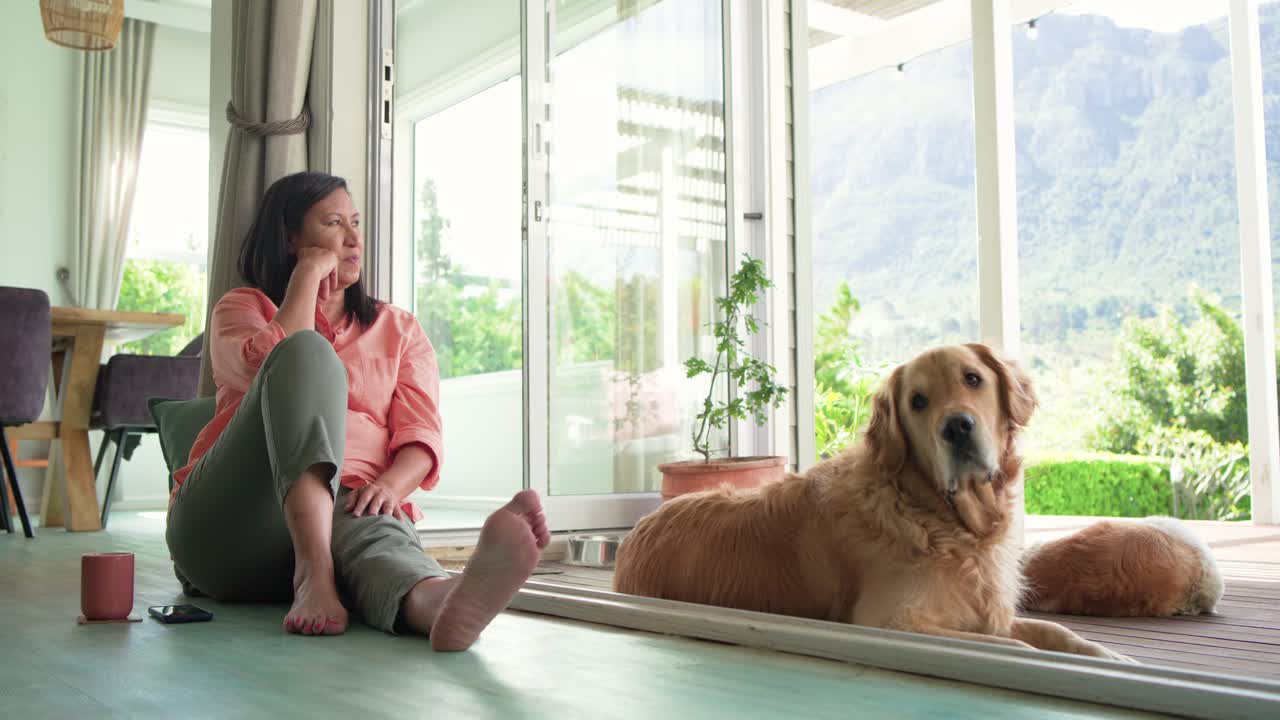 Una donna matura, seduta per terra accanto a una porta scorrevole, osserva il panorama dopo che un golden retriever ha mosso la testa.
