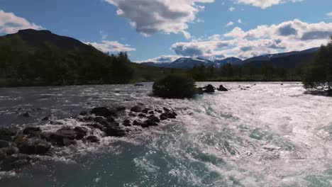 sun reflecting off the surface of a wide river flowing through a lush green forest in norway on a beautiful summer day. aerial push forward shot