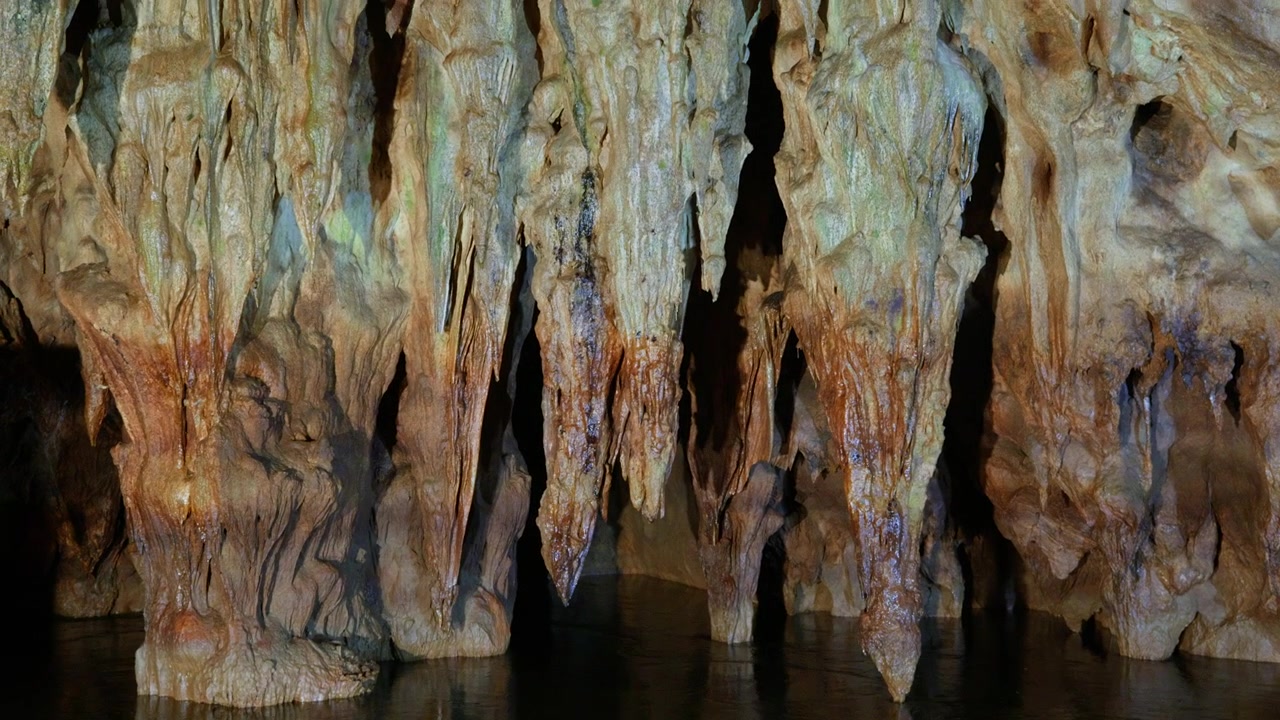 Multiple limestone stalactites with orange and white mineral deposits hang from cave ceiling above dark underground water. Slow lateral pan right reveals intricate rock formations and textured.