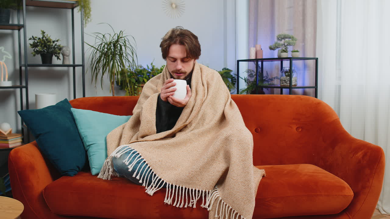 Sick ill caucasian man wrapped in plaid sit shivering from cold drinking hot coffee or tea at home