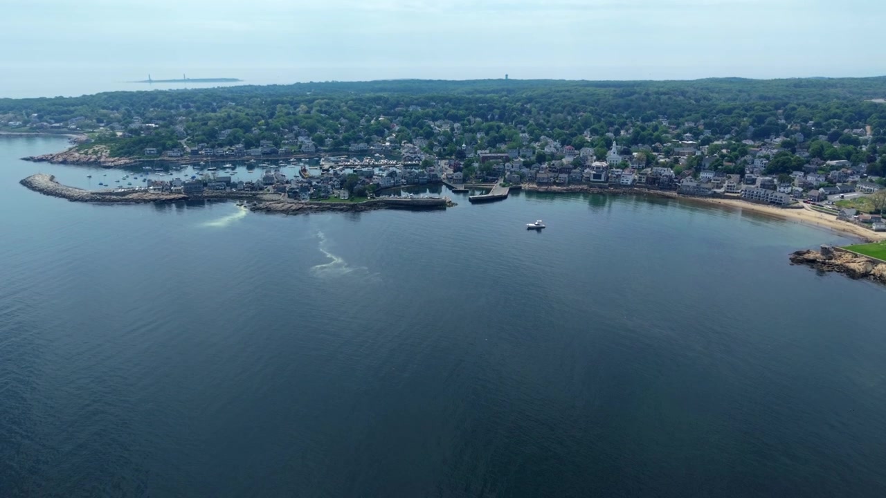 Bahía de Rockport, Massachusetts, con pueblo portuario, costa de granito, playa de arena y estela de barco en aguas tranquilas, avance aéreo y picado.