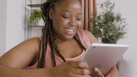 close up view of woman with braids lying on a big sofa while smiling and using a tablet