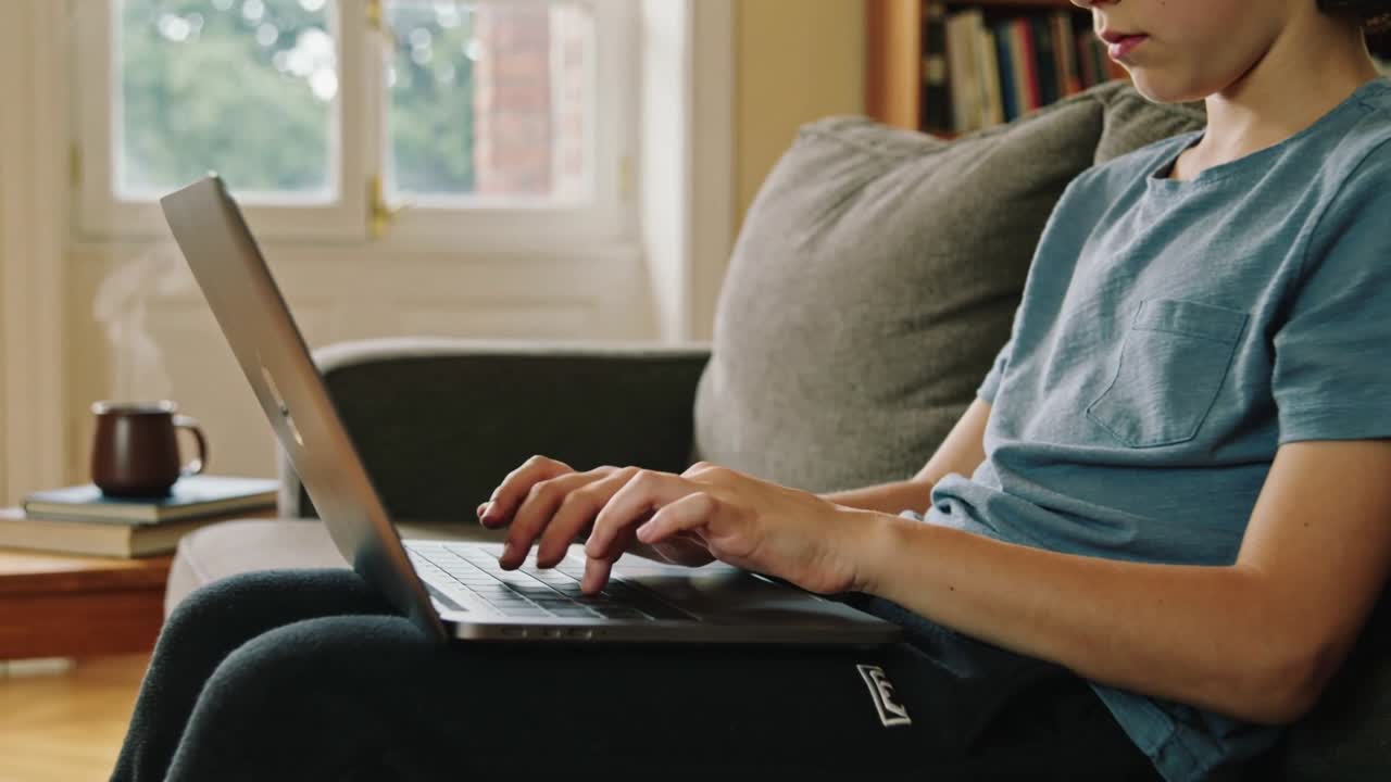 Settling back, teen male typing on laptop on sofa, studying for school with steaming mug