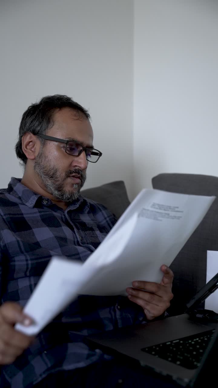 Homem Lendo Papéis, Organizando Informações Financeiras Enquanto Sentado em um Sofá. Vídeo Vertical