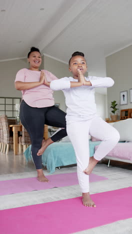 vertical video of happy african american mother and daughter doing yoga at home, in slow motion