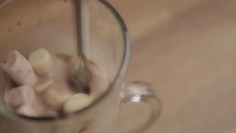 stirring glass mug of hot chocolate with marshmallows and whisk