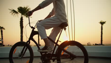 Ein-Glücklicher-Mann-In-Weißen-Hosen-Tritt-In-Die-Pedale-Und-Fährt-Mit-Seinem-Schwarzen-Fahrrad-Am-Morgenstrand-Entlang-Des-Meeres-Bei-Sonnenaufgang-Im-Sommer