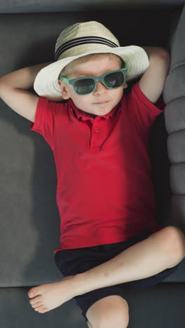 relaxed toddler in sunglasses enjoys lying on sofa near suitcase after arriving. boy in straw hat takes break in hotel room upper close view