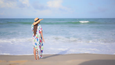 mujer solitaria y elegante con colorido vestido de verano y sombrero de pie en la arena frente a las olas del mar tropical, vista trasera