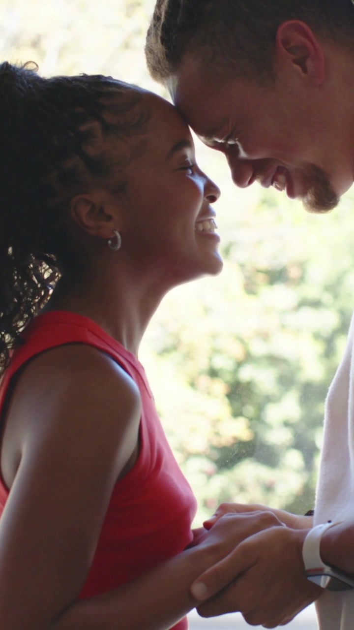 Vertical video: Holding hands pair bonding leaning and touching foreheads on porch wearing red top