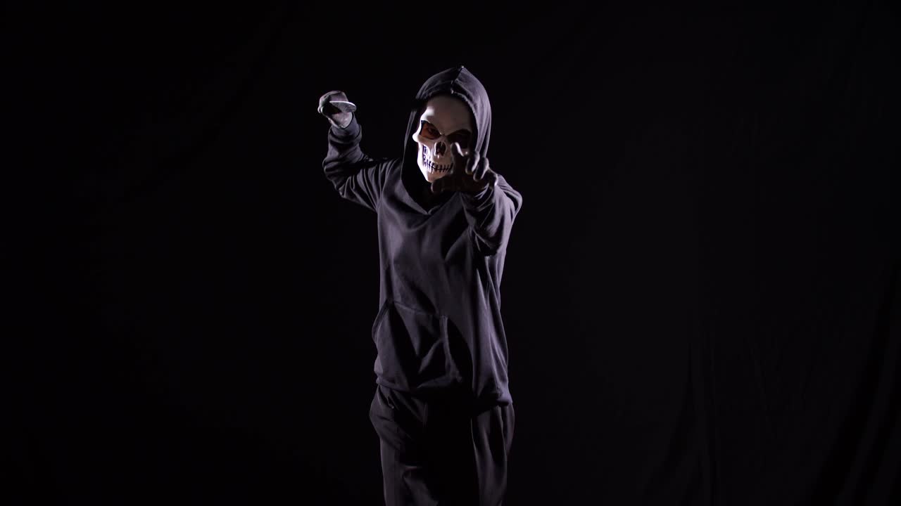 страшный мужчина в пуловерке с капюшоном, носящий маску хэллоуина, держащий нож и указывающий на камеру во время бега на черном фоне