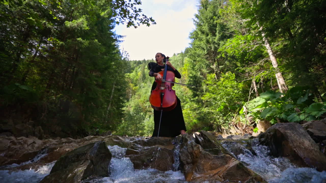 Luxury woman with musical instrument outdoors. Female musician performing music with a cello on amazing nature background with fresh mountain water.