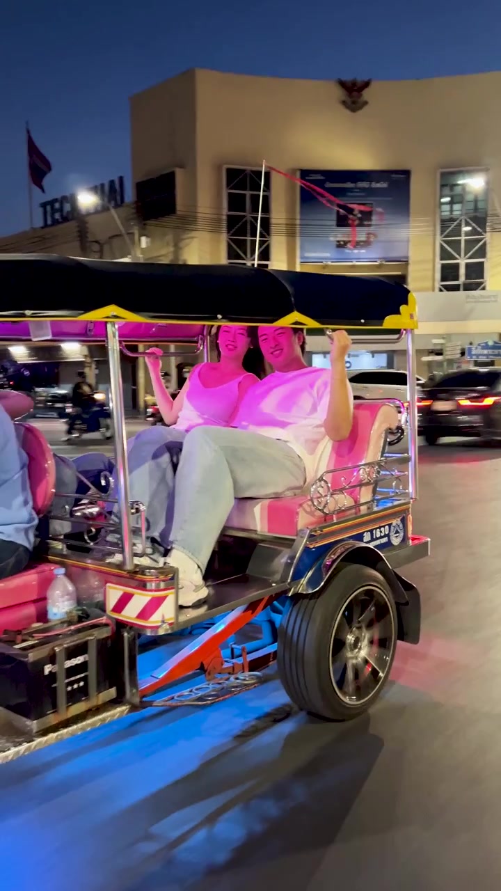 Casal sorri e relaxa em tuk-tuk colorido, luzes da cidade brilhando, câmera acompanhando o veículo.