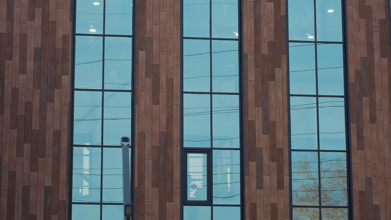 Glass facade of modern fitness center reflects outdoor trees while soft lights illuminate interior space, with subtle movement of figures visible through clear blue-tinted vertical windows