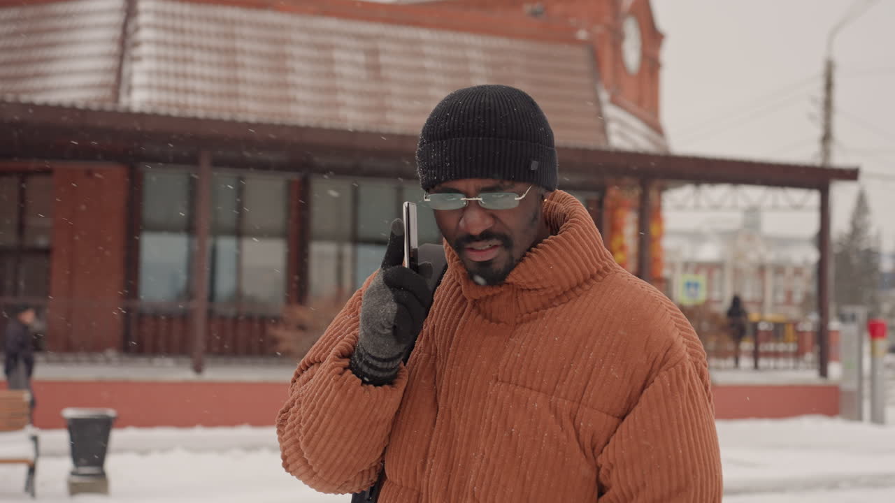 Uomo di colore con cappotto arancione che parla al suo smartphone fuori da un caffè in mattoni durante una leggera nevicata, indossa berretto, guanti e zaino, pendolare calmo che controlla le indicazioni, luce urbana soffusa, calda texture di lana