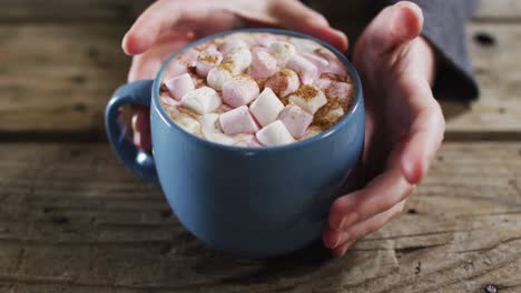 close up view of hands holding a hot chocolate with marshmallows against wooden surface