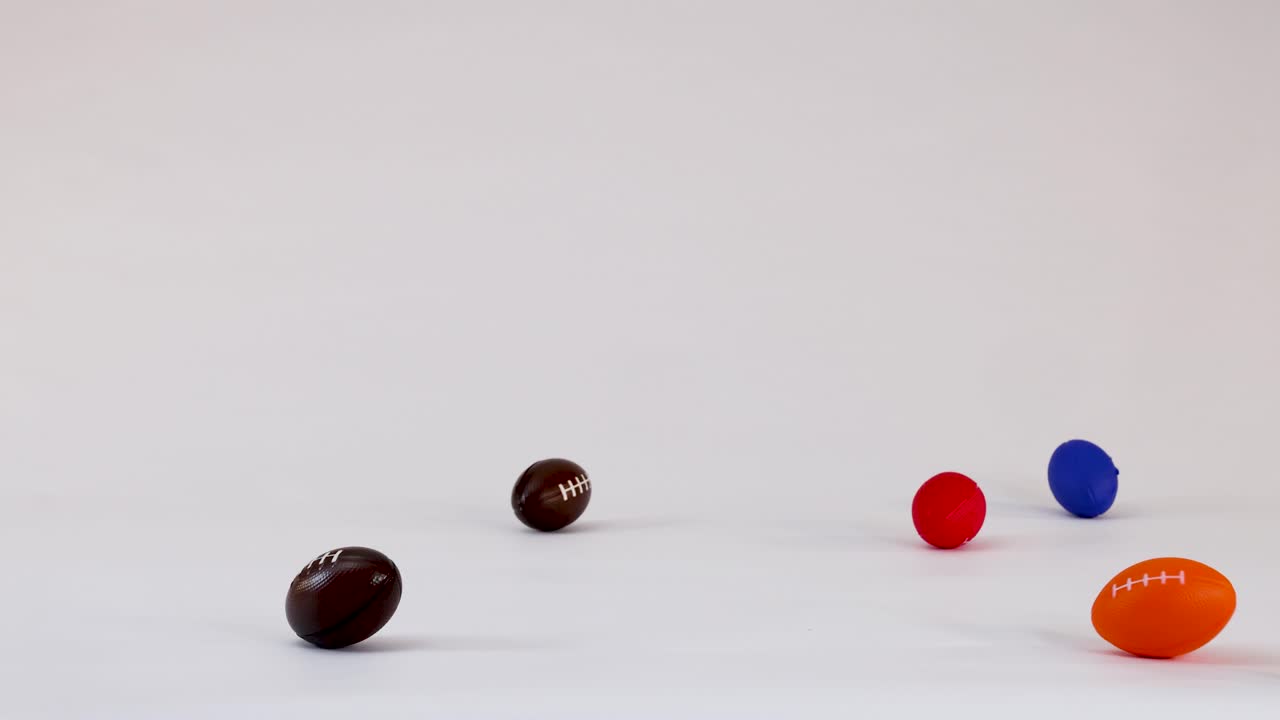 Multiple small colorful foam footballs fall, bounce, and roll across a plain white surface