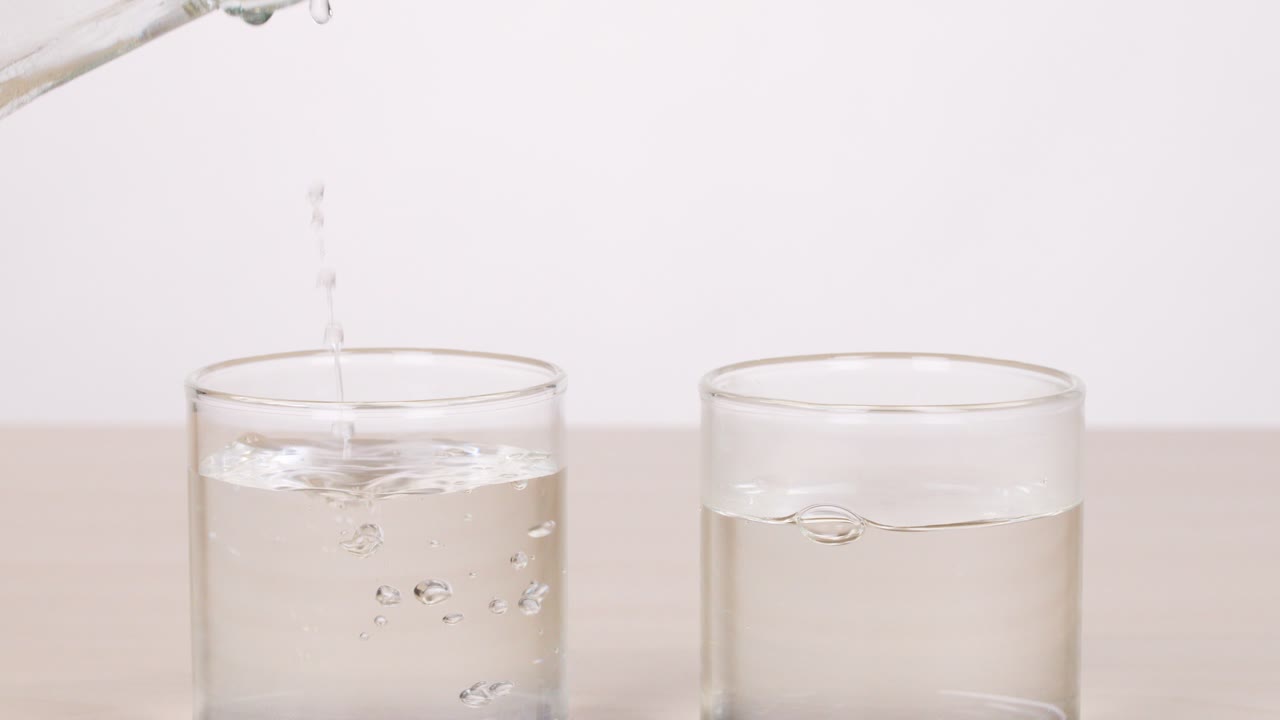 Clear water poured from glass bottle into two glasses, neutral background, steady camera, bright lighting