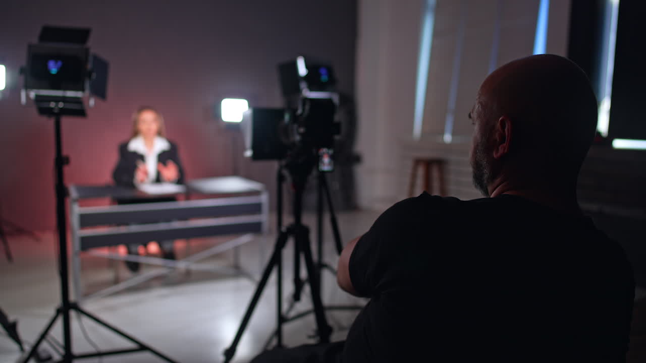 Rear view of a male photographer sitting backstage in front of camera. Unrecognized long-haired woman in black jacket sits at desk talking.