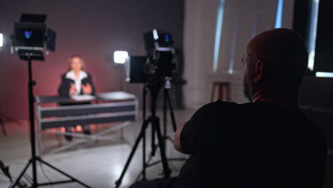 rear view of a male photographer sitting backstage in front of camera. unrecognized long-haired woman in black jacket sits at desk talking.