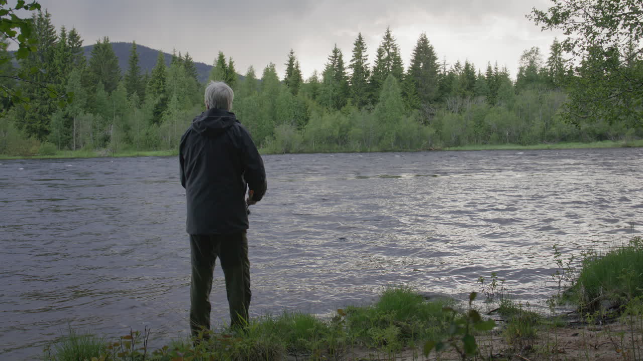 Older man standing by a riverbank fishing in the calm flowing water surrounded by forest and distant hills. Filmed at Glomma river, in Norway