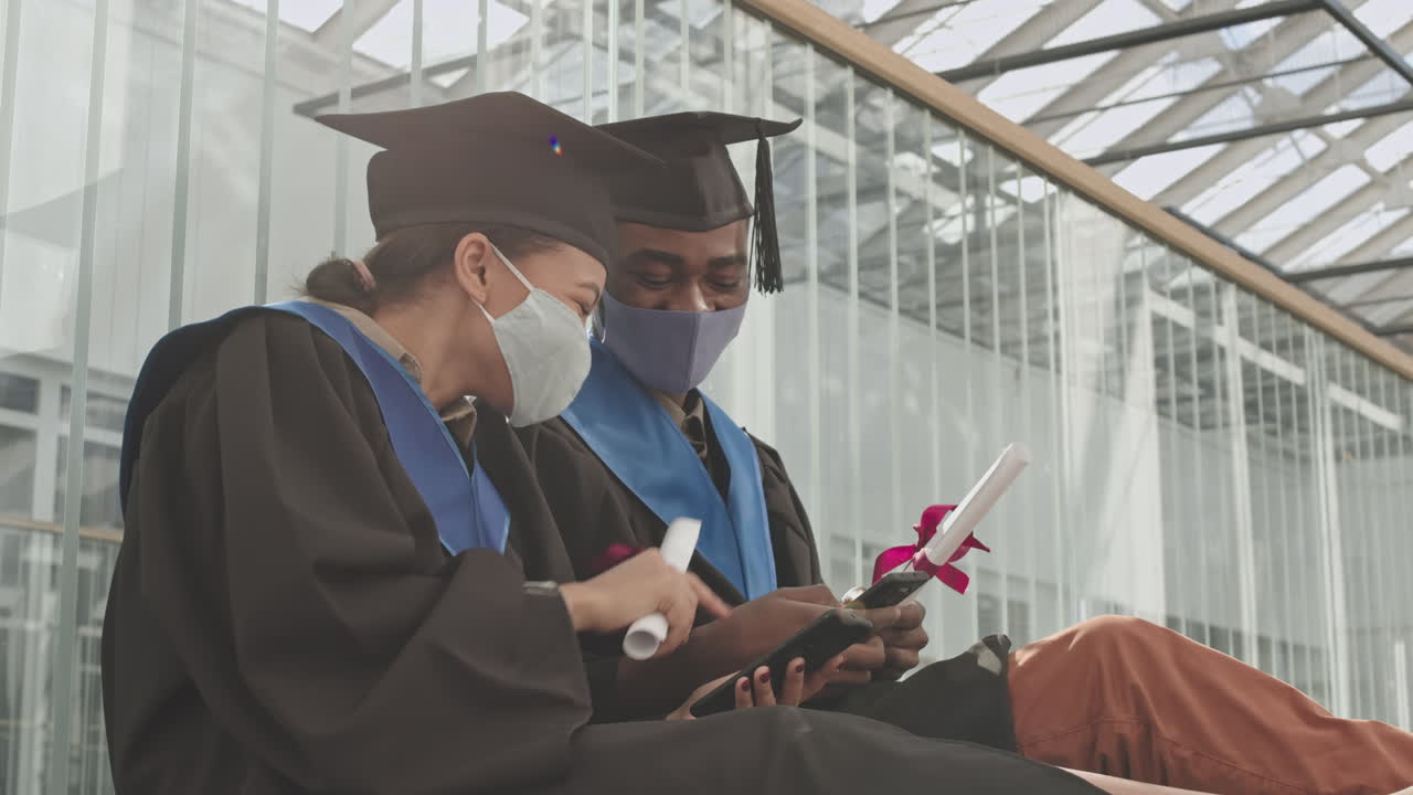 Two Young Multiethnic Graduates Using Cellphones