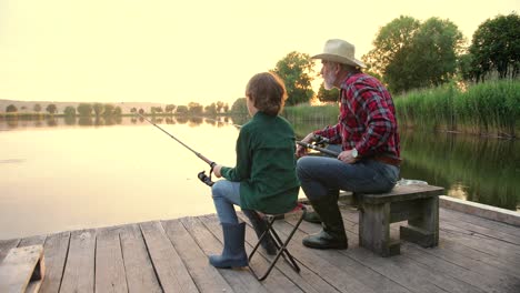 camera zooming on rear view of a teen boy sitting with his grandfather on the lake pier, talking and fishing together