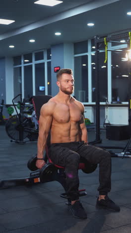 handsome athlete training with dumbbells. shirtless sportsman with muscular body lifting heavy dumbbells to the sides while sitting in the modern sports club. vertical video