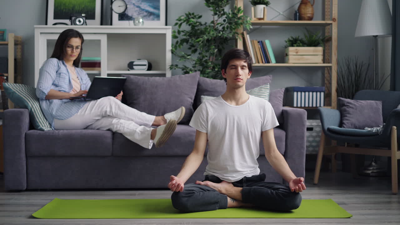 Couple relaxing at home: woman working on laptop, man meditating