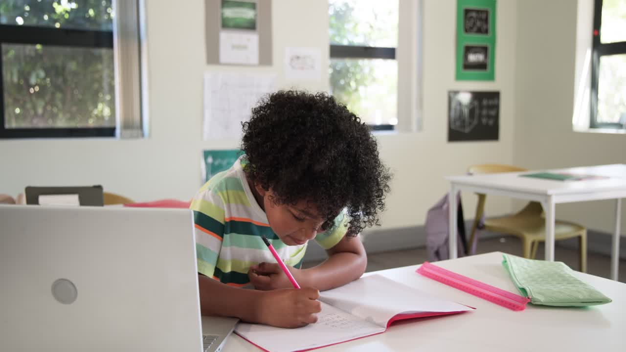 Student is writing in notebook at desk, checking laptop for instructions as camera moves in