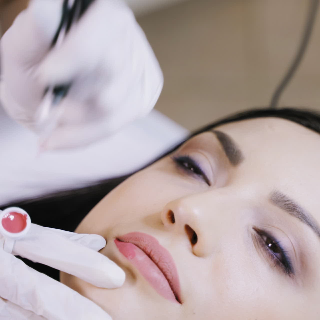 A beautician gently applies a pink hue pigment to the client's lips using a professional permanent makeup machine. Close-up.