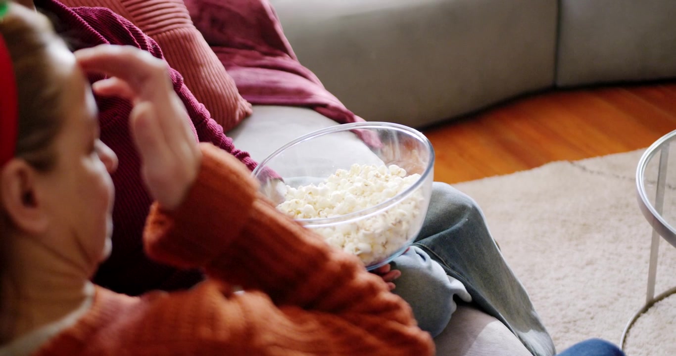 Amiche con maglie color ruggine e bordeaux che condividono una ciotola di popcorn in vetro sul divano, la donna a sinistra si sistema i capelli