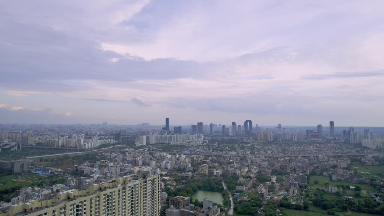 Plano aéreo com drone voando para trás, mostrando edifícios de vários andares da cidade se estendendo para longe e coloridas nuvens de monção acima