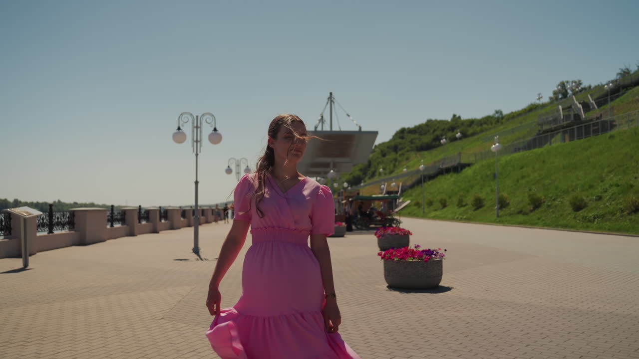 Mulher de vestido rosa caminhando pelo calçadão à beira-mar com postes de luz e floreiras, luz ensolarada do meio-dia, passarela de paralelepípedos, brisa suave levanta a saia, enquadramento cinematográfico, clima romântico e tranquilo