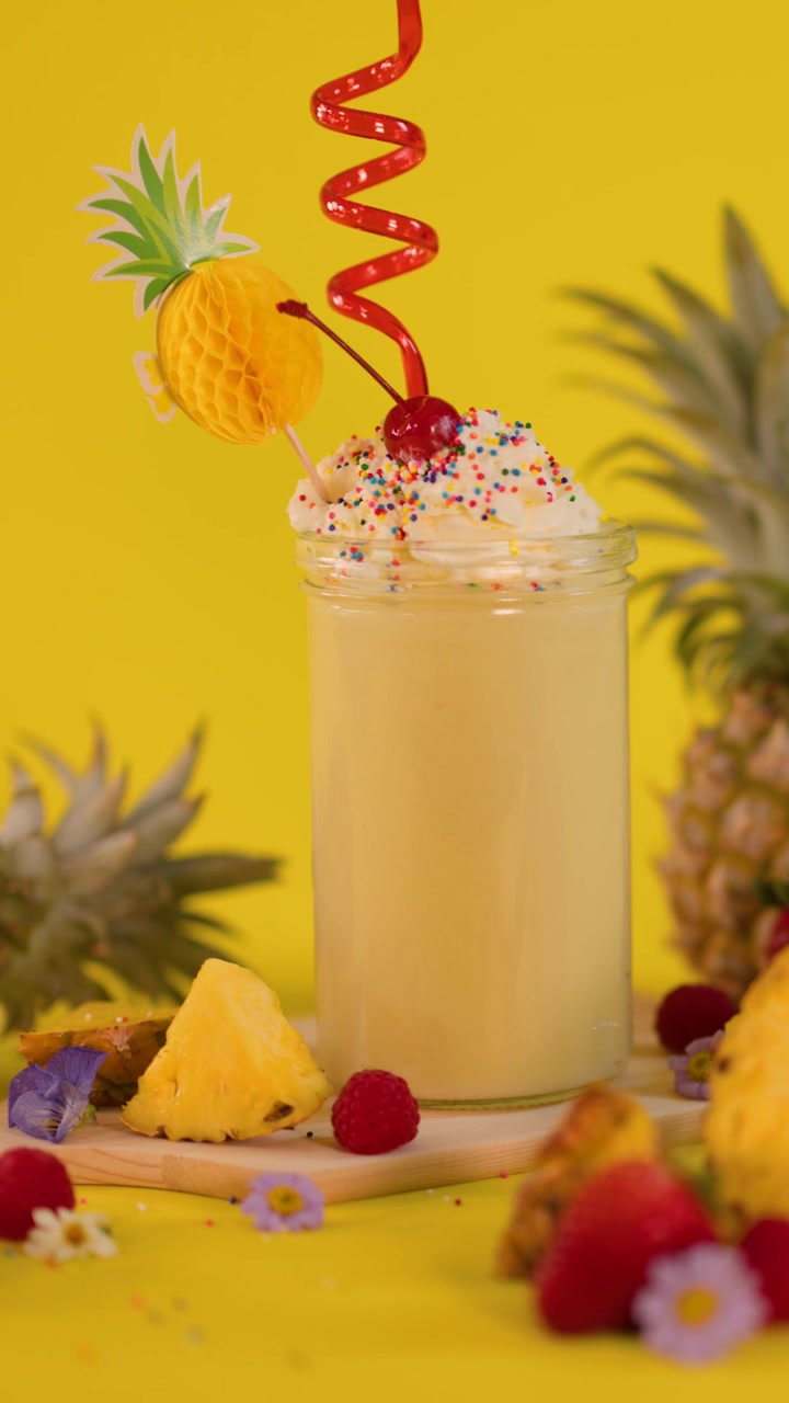 Una mano aggiunge una cannuccia rossa riccia e festosa a un frullato all'ananas su sfondo giallo