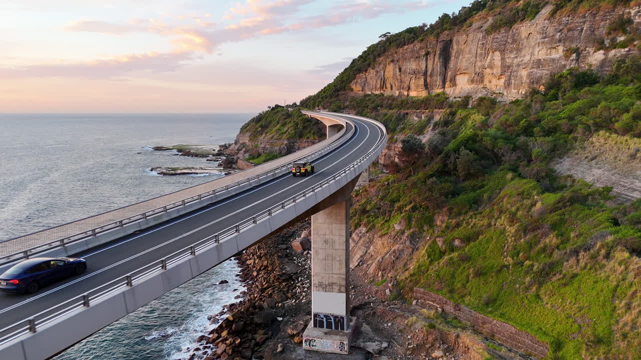 Aerial tracking footage of a car along the Sea Cliff Bridge, highlighting the breathtaking road suspended over the sea. Cinematic drone footage.
