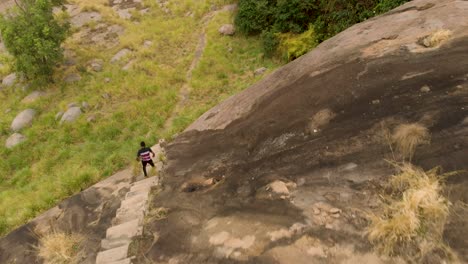 Toma-Aérea-Vista-De-Pájaro-De-Un-Hombre-Africano-Bajando-Las-Escaleras-Al-Lado-De-Una-Montaña-De-Granito