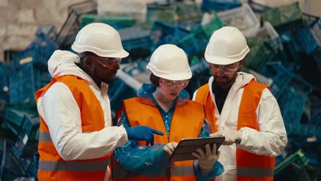 A-brunette-girl-in-a-blue-uniform-and-a-White-protective-helmet-in-an-orange-vest-holds-a-tablet-in-her-hands-and-tells-her-fellow-guys-about-her-plans-at-the-plastic-and-waste-recycling-plant