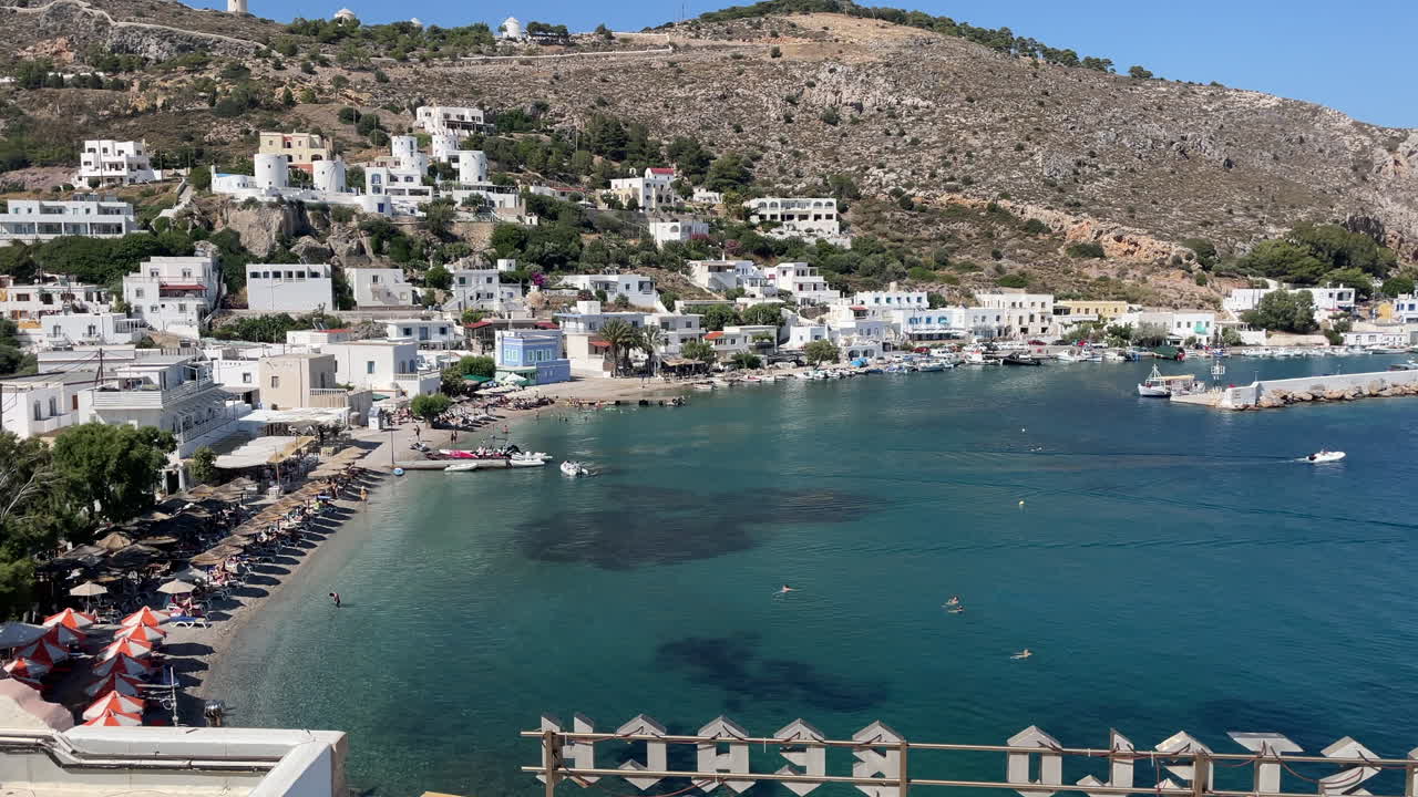 Greece,Leros,Panteli Bay, fishing port, Panteli village and beach on a sunny day. Boats anchored at the bay, some people swimming, some sun bathing, white houses at the slope, castle and windmills.