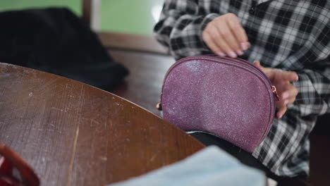 closeup view of young woman sitting on wooden chair, picking up sparkly purple cosmetic bag from wooden table, and removing beauty tools, elegant nails and soft lighting add to the aesthetic